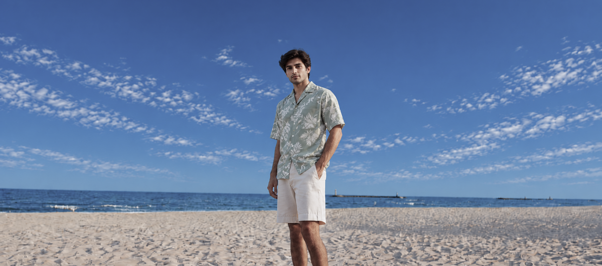 A man stands on a sandy beach wearing a light green tropical print short-sleeve button-down shirt and off-white shorts, with the ocean and blue sky behind him.