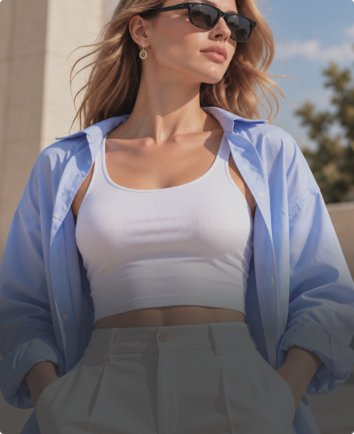 A woman wearing a white ribbed tank top, an oversized light blue button-up shirt, and light gray trousers, photographed outdoors with sunglasses and a small hoop earring visible.